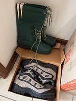 Tall green NFL Green Bay Packers boots next to a box containing gray and black hiking boots, both pairs new.