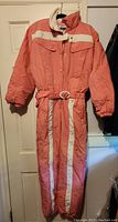 Coral pink padded women's vintage ski suit with white stripes, belt, and snap buttons on hanger