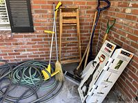 Photo showing yard tools including garden shovels, seed spreader, broom, garden hoses coiled on floor against brick wall