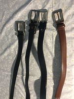 Four men's belts laid out on a surface, three black and one brown, each with silver buckles, showing full length.