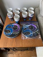 Top-down view of multiple stacks of 9-inch paper plates and paper cups arranged on a wooden table. Several packs of unopened plates and cups displayed, all feature a colorful '50... the party continues' design on dark background.