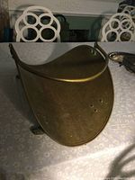 Antique brass wood holder with curved body and handle. Shown from an angled front view on a table with dining chairs in the background.