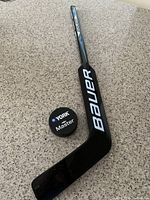Mini Bauer hockey stick alongside York Master black hockey puck on a speckled countertop, displaying stick blade and side profile.