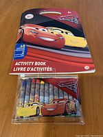 Image showing the front cover of the Cars activity book featuring Lightning McQueen and Cruz Ramirez with a set of 8 crayons wrapped in Cars character paper under the book.