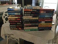 Photo shows a collection of 34 hardcover books displayed on a lace-covered table. Books are primarily by James Patterson, arranged in three stacks with spines facing outward for title visibility.