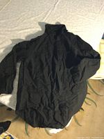 Photo of a black long sleeve linen-cotton shirt laid flat on a light surface. The shirt features an asymmetrical cut with a longer back and a high collar.