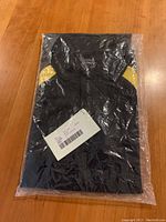 Top view of the folded black polo shirt in its plastic retail packaging with attached product tag.