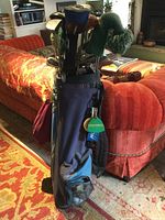 Full view of dark blue golf bag with multiple golf clubs inside, standing against a red sofa in a living room setting.