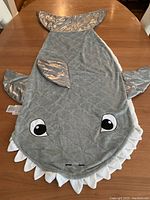 Top-down view of the shark-shaped blanket sleeping bag laid flat on a wooden table showing detailed features like the shark eyes, white teeth edging, and shiny silver fins and tail.