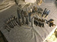 Photo of assorted specialty cutlery spread on table including spoons, forks, knives, and cutting tools, mostly metal with some mother of pearl handled pieces