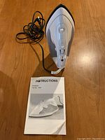Top view of white steam iron with instruction manual placed below, showing iron heating surface and buttons.