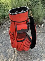 Side view of an orange Titleist golf bag with black trim, padded strap and multiple pockets.