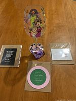 Photo showing hand painted wine glass with colorful design of a woman and decorative base next to three individually packaged fridge magnets with wine-themed quotes.