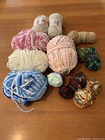 Photo showing twelve yarn skeins and ends arranged on a wooden floor, highlighting different fiber textures and colors including chenille, cotton blend, and variegated yarns.