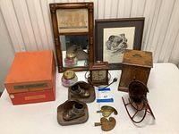 Photo shows a collection of antique decor on a white table including framed mirror with ship print, framed dog art, Seth Thomas plug-in clock, wooden storage box, bronzed baby shoe bookends, a clear glass container with lid, boxed puzzles, and small vintage sleigh decor.