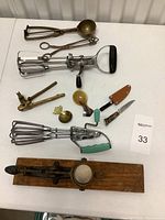 Varied antique kitchen utensils arranged on a table showing egg beaters, ice cream scooper, nutcracker, salad tongs, and small spoons and knives