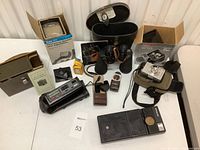 Full view of lot items including cameras, binoculars, slide viewer, alarm clocks and accessories arranged on a white surface