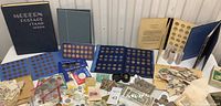 Wide view of two stamp albums, multiple stamp and coin stock pages, loose coins in tubes and assorted world currency laid out on table