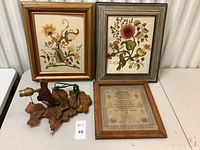 Grouped view of two framed floral cross-stitch artworks and one framed cross-stitch almanac with rustic wooden lamp base on table