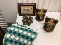 Full view of the lot on white table showing 6-piece silver plated Greek egg cruet set, bison bookends, framed fabric horse race art print, and green crocheted blanket