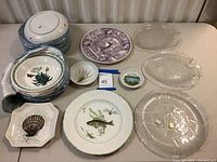 A mixed collection showing stacked dishware, plates with botanical and fish designs, a purple StaffordshireWare plate, and various clear glass fish plates.