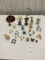 Collection of 21 varied metal egg stands in gold and silver tones arranged on white surface, showing details and wooden bases on largest stands.