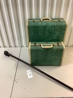 Two stacked green Samsonite luggage pieces with gold-tone handles and latches beside a dark brown carved wooden cane lying on the table.