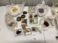 Multiple mineral specimens displayed on a table, including amethyst clusters, calcite, and wulfenite crystals, some wrapped in tissue paper or displayed in small white boxes.