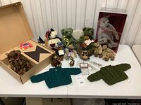 Overview of teddy bear collection including grey Steiff bear in case, assorted small bears, crocheted sweaters, eyeglasses, and other accessories on table.