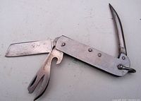 Photo showing multi-tool partially open with visible marlin spike, blade, and hooked forked prong tool, metal surface showing signs of wear and patina. Tool length approx 4 inches.