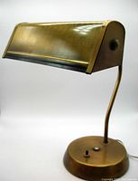 Front-right angle view of vintage banker desk lamp showing rectangular hinged shade and curved neck with antique brass finish.