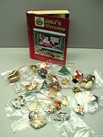 Photo of the book-shaped tin labeled 'Santa's Workshop' alongside 25 Christmas-themed brooches individually sealed in plastic bags scattered in front of it on a flat surface.