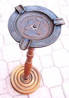 Full view of floor ash tray showing cast iron top with symbols and wooden stand base on paving stone ground.
