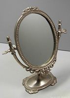 Front angle view of the vintage heavy adjustable oval vanity mirror showing ornate metal frame and base with embossed decorative details.