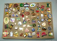Top view of a textured beige backing board displaying about 70 assorted vintage pins featuring various logos, figures, and commemorative designs.