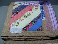 Folded large patchwork quilt with multi-color diagonal fabric strips and tan floral border.
