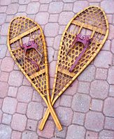 Pair of vintage rawhide snowshoes laid crossed on a brick floor, showing the front side with leather bindings and the wooden frame.