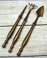 Three solid brass indoor garden tools laying parallel on a wood patterned surface showing the full length and design.