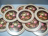 Set of twelve round placemats stacked together showing floral basket design with roses on a dark brown background.