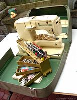 Vintage Bernina Record 730 sewing machine in off-white color placed inside its open green hard-shell carrying case. The multi-tiered golden-yellow accessory box containing bobbins with colorful thread, screwdrivers, and various metal attachments is positioned in front of the machine.