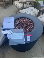 Round black gas fire pit filled with reddish-brown lava rocks with instruction manuals and documents placed on the side.