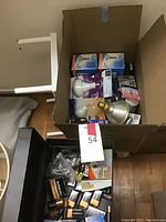Box containing multiple packs of Duracell and Energizer batteries alongside various lightbulbs, a silver metal lamp shade, and lamp components.