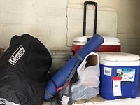 Wide shot showing Coleman inflatable mattress in carry bag, blue and red coolers, rolled blue sleeping mat, and additional camping gear bags