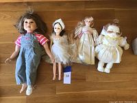 Four dolls lying on wooden floor: large girl doll in denim overalls, ballerina doll, seated doll in pink dress, white fluffy doll
