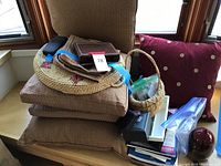 Stack of four brown fabric pillows with geometric and polka dot prints. Woven basket tray on top with a blue ribbon. Dark eyeglasses case beside them.