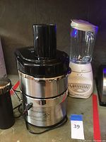 Front and side view of Jack Lalanne Power Juicer showing stainless steel body, black plastic feed chute, power cord, and red switch. Also showing black blender container with silver lid beside it.