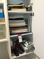 View of paper products and notebooks stacked inside a white cabinet shelf with additional office tools in a plastic basket below