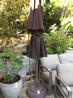 Full view of the umbrella stand with closed fabric umbrella, placed outdoors near potted plants and patio chairs.