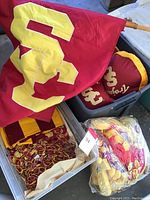 Photo showing a large red and yellow USC flag, red and yellow pom poms in plastic, and fabric items with USC logos and Trojan text in bins.
