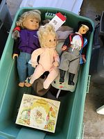 Three vintage dolls and a Beatrix Potter children's tea set box inside a green bin.
